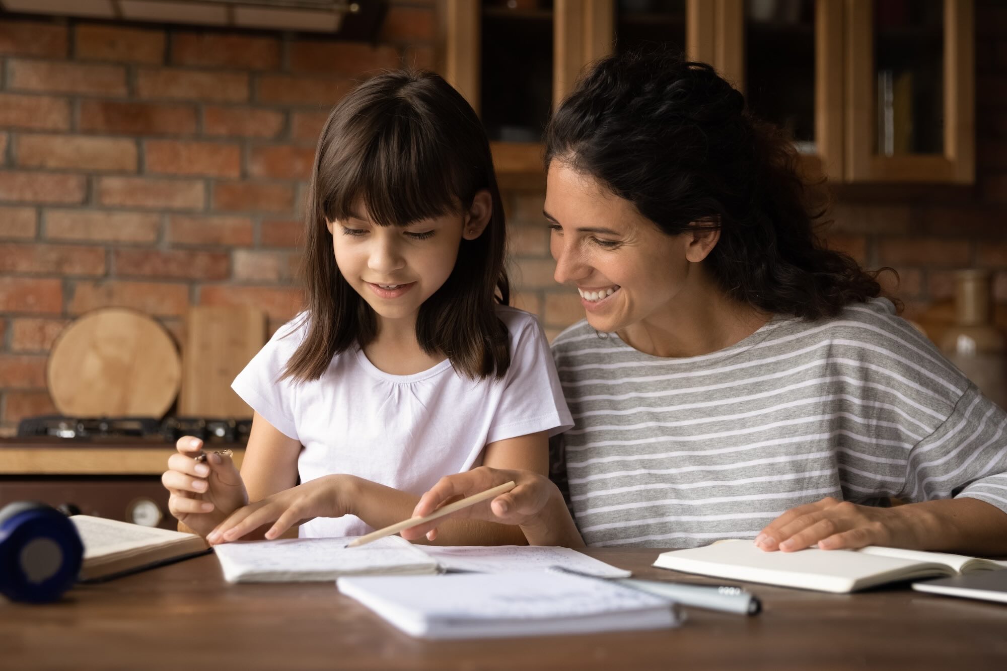 Little girl smiling working on school work next to her mother who is smiling wearing striped shirt