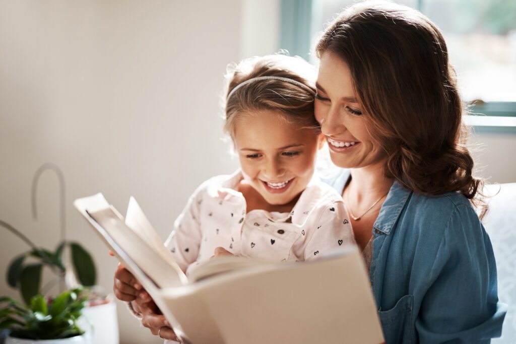 A mother and young daughter sit together indoors, smiling while reading a book, with soft natural light coming through a nearby window.