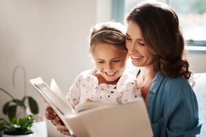 A mother and young daughter sit together indoors, smiling while reading a book, with soft natural light coming through a nearby window.