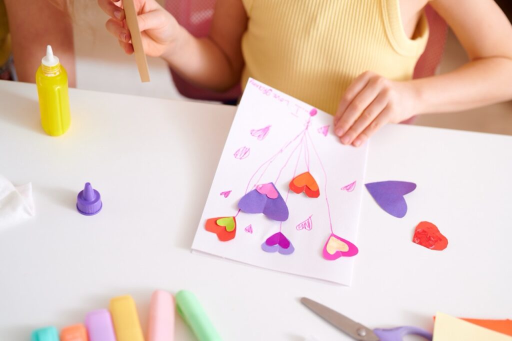 Child making a Valentine’s Day card with colorful paper hearts, glue, and craft supplies at a table.