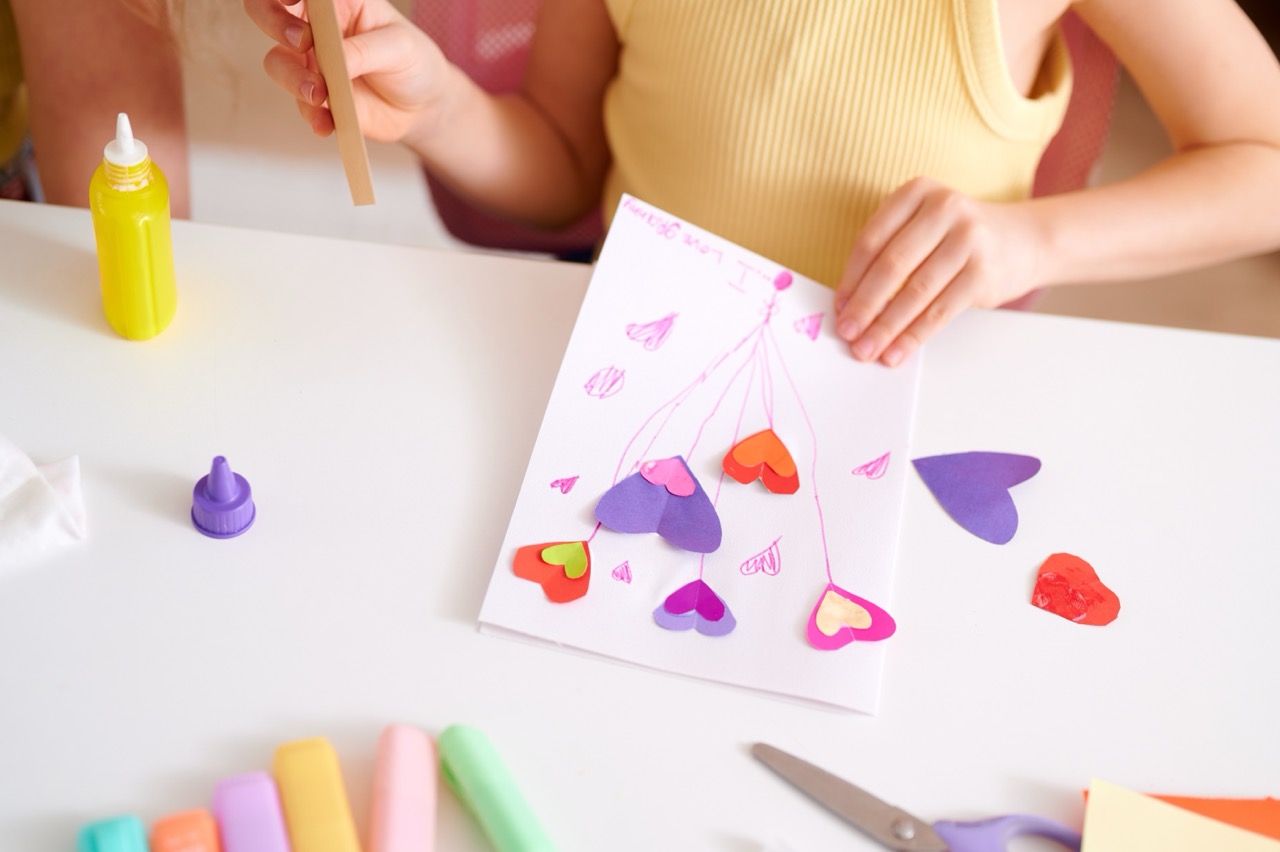 Child making a Valentine’s Day card with colorful paper hearts, glue, and craft supplies at a table.
