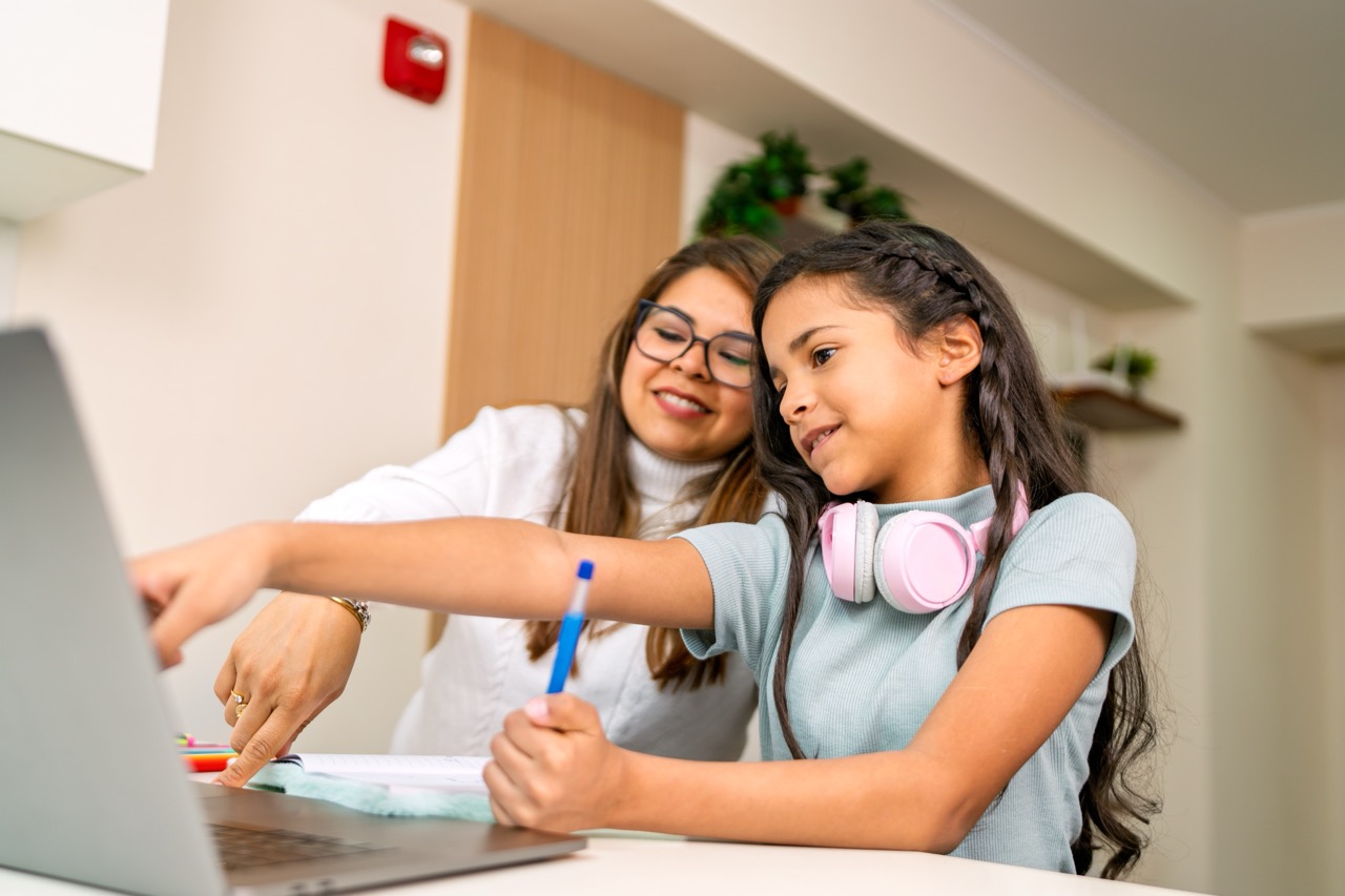 A mother and young daughter sitting together at a laptop, reviewing schoolwork at home in order to finish the year strong, with the girl holding a pencil and wearing pink headphones around her neck.
