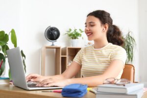 Smiling young woman engaged in remote learning or online work at home desk with laptop and earbuds, she is using a laptop computer and wearing wireless earbuds