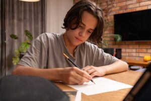 Focused teenage boy writing or drawing on paper with pencil at wooden table. Young male student doing homework or sketching in cozy home interior with brick wall and indoor green plants.